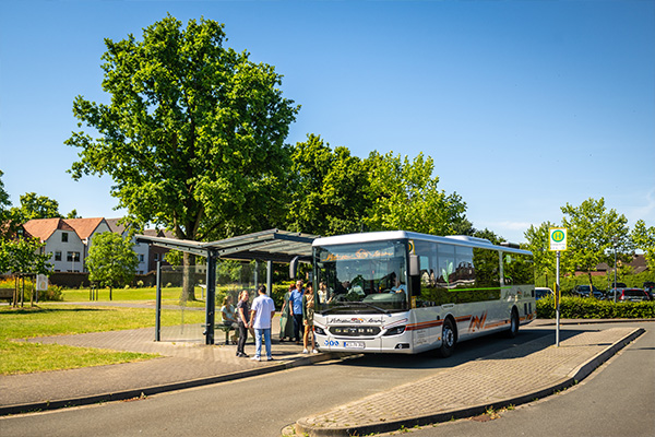 Ein silberner Linienbus der Firma Verhuven steht an einer Bushaltestelle, an der Leute auf den Bus warten