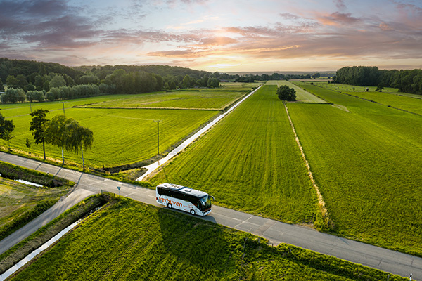 Zwischen grünen Feldern und bei Sonnenuntergang ist in der Entfernung ein weißer Verhuven Reisebus zu sehen, der über die Landstraße fährt.