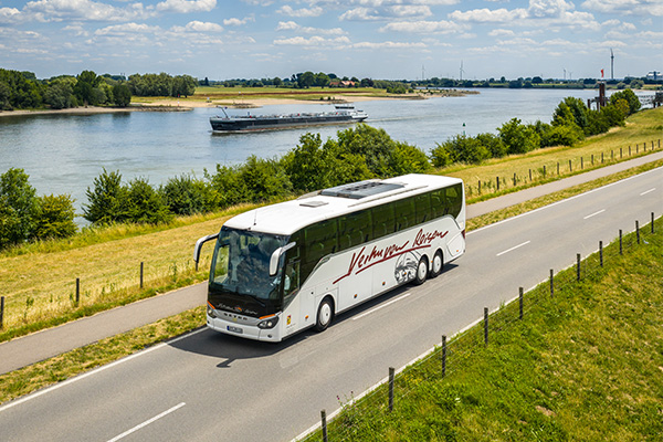 Ein moderner Reisebus mit der Aufschrift Verhuven Reisen fährt durch eine grüne Landschaft.